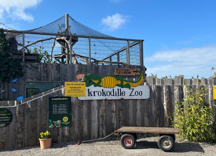Indgangen til Krokodille Zoo med et træhegn, et skilt med en tegnet krokodille og informationstavler om dagens program. Foran hegnet står en lille trækvogn, og bagved ses et indhegnet anlæg under blå 
