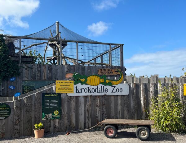 Indgangen til Krokodille Zoo med et træhegn, et skilt med en tegnet krokodille og informationstavler om dagens program. Foran hegnet står en lille trækvogn, og bagved ses et indhegnet anlæg under blå 