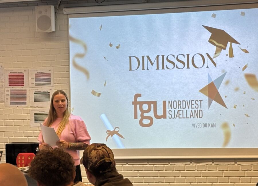 En medarbejder holder tale ved en dimission foran en storskærm med FGU Nordvestsjællands logo, mens elever sidder og lytter.