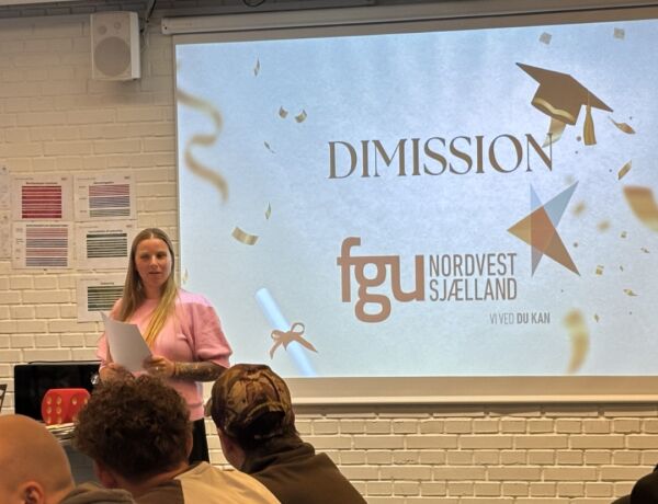 En medarbejder holder tale ved en dimission foran en storskærm med FGU Nordvestsjællands logo, mens elever sidder og lytter.