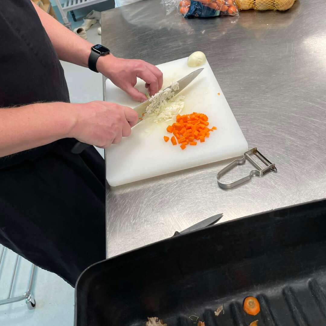 Person hakker løg og gulerødder på et skærebræt i et køkken.