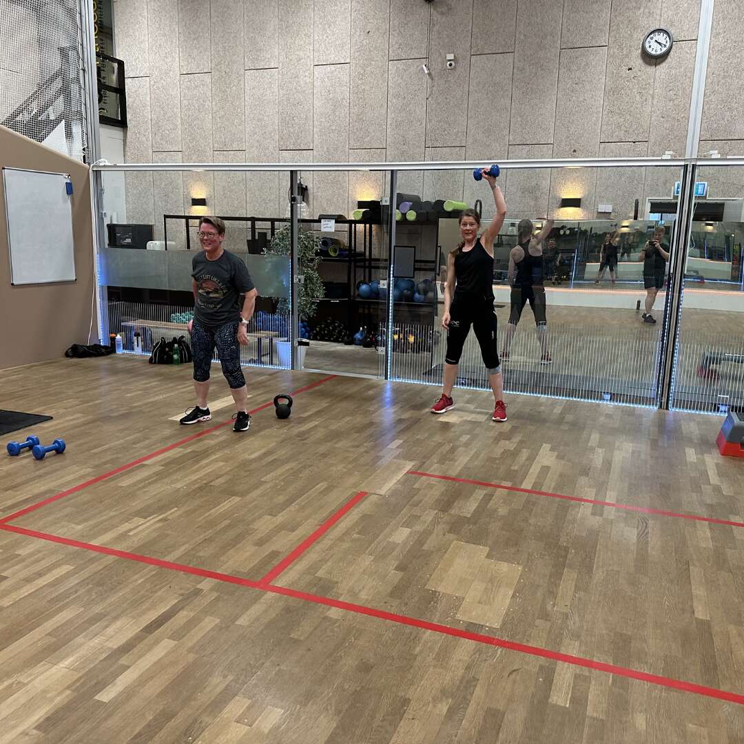 To personer træner med håndvægte og kettlebell i et fitnesslokale.